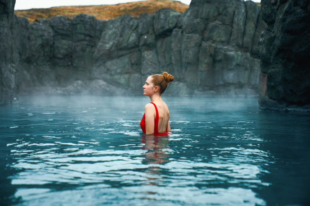 Girl in red swimsuit stands in the misty water of Sky Lagoon, surrounded by dark rocks.