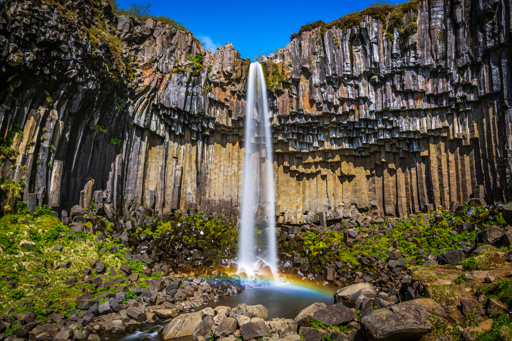 Svartifoss waterfall cascading down from basalt columns to form a rainbow in a pool in South Iceland.