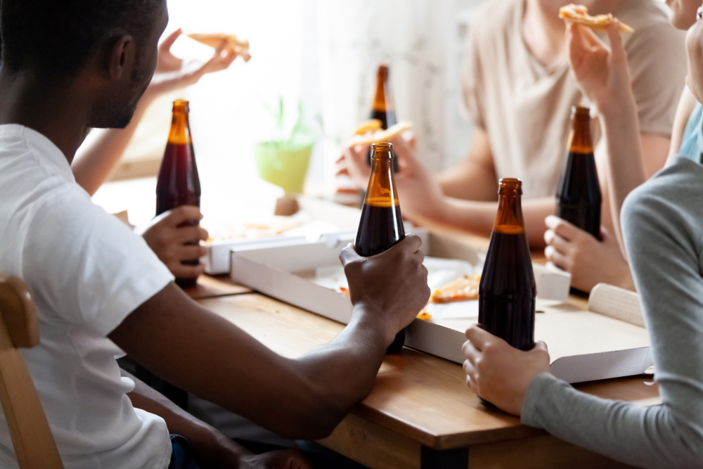 Close up mixed race group of cheerful people sitting at served table, holding glass bottles and drinking beer, eating  pizza from cardboard box,