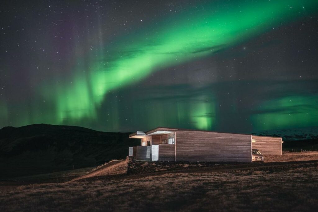 A wooden building on the side of a hill with mountains in the distance and the green northern lights in the sky. 