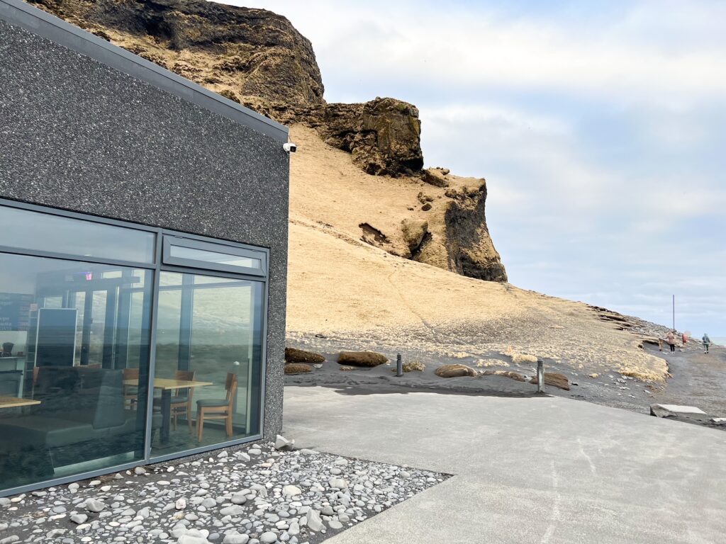 Corner of a black cafe by the black beach with a sandy cliff behing the cafe. The cafe has large windows  