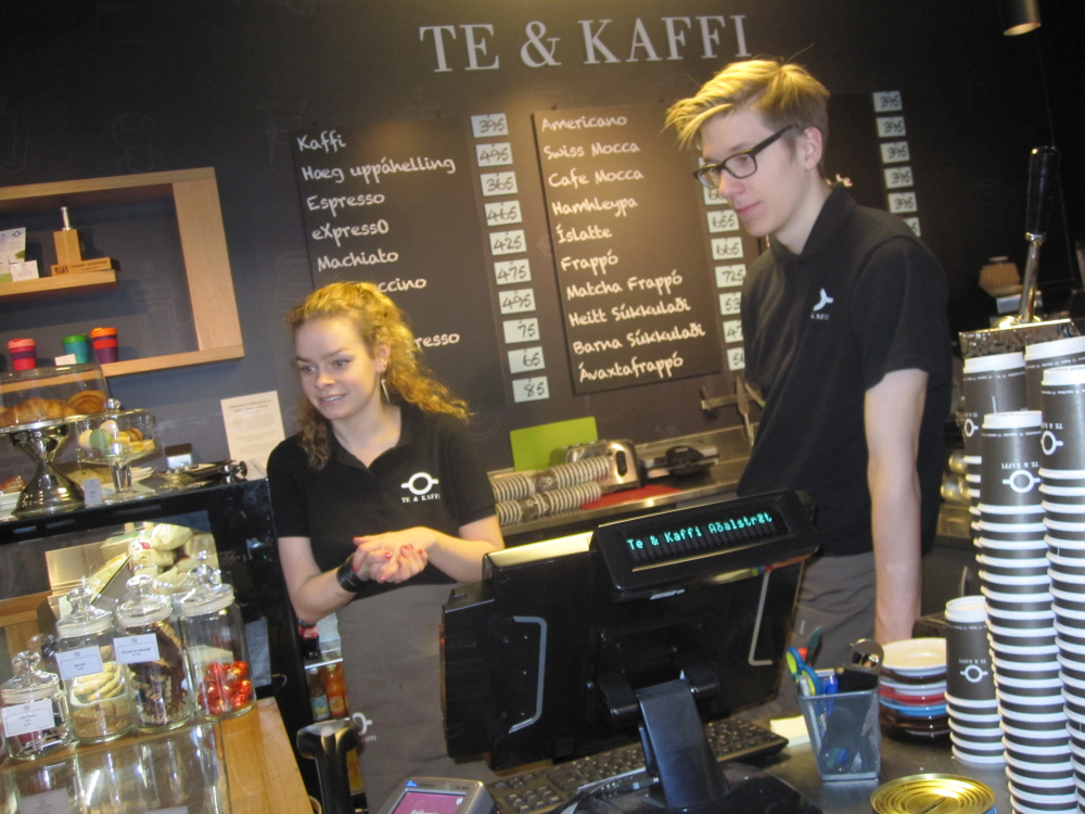 Coffee shop interior of Te & Kaffi  one of the places for coffee in Iceland. There are two servers behind the counter and a black menu board in the background. 