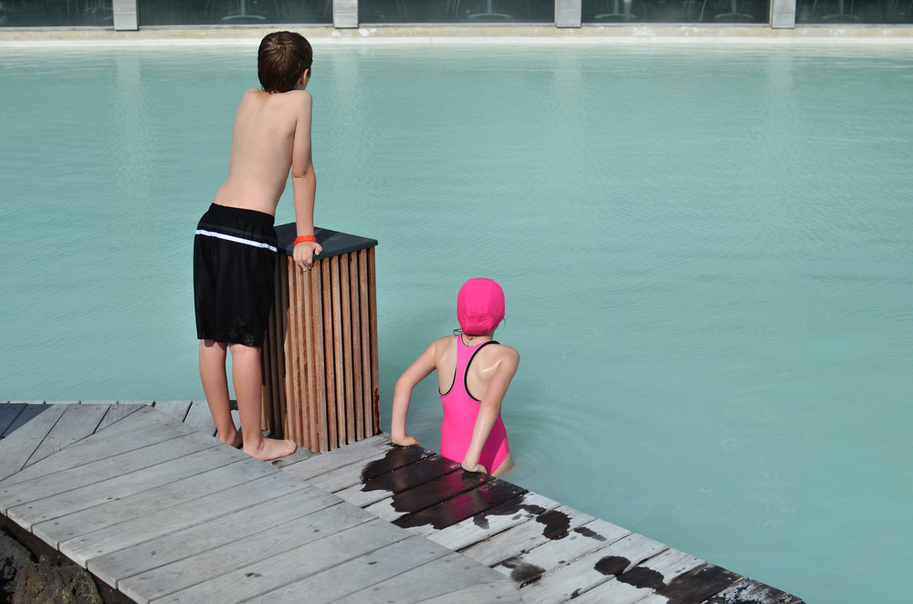 Two children in a swimming pool on is stool on the side. 