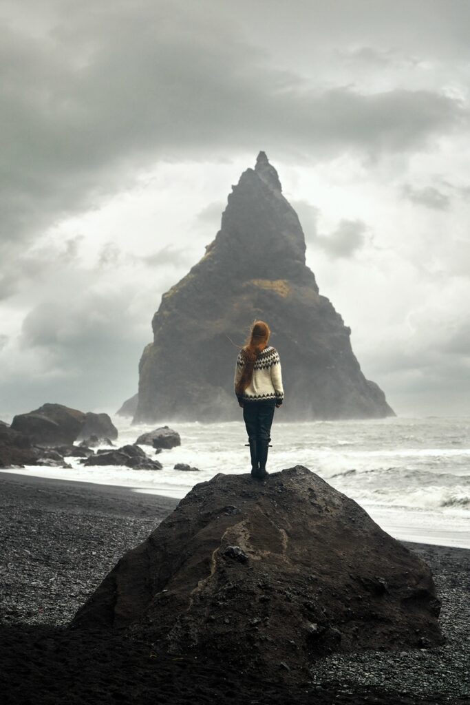 A woman standing on a rock on a black sand beach wearing an Icelandic sweater. 