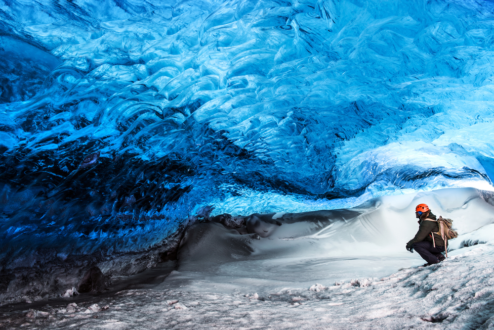 A person crouches below the blue light of an ice cave during one of the Iceland south coast tours. 