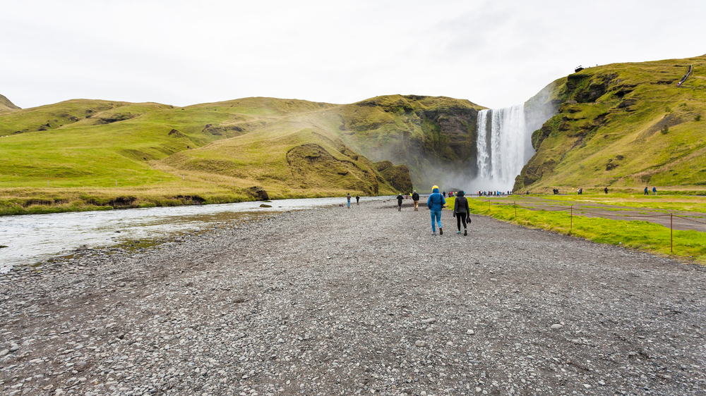 A group of tourists approach a gigantic waterfall on one of those Iceland South Coast tours: they walk toward the falls on a gravel road. 