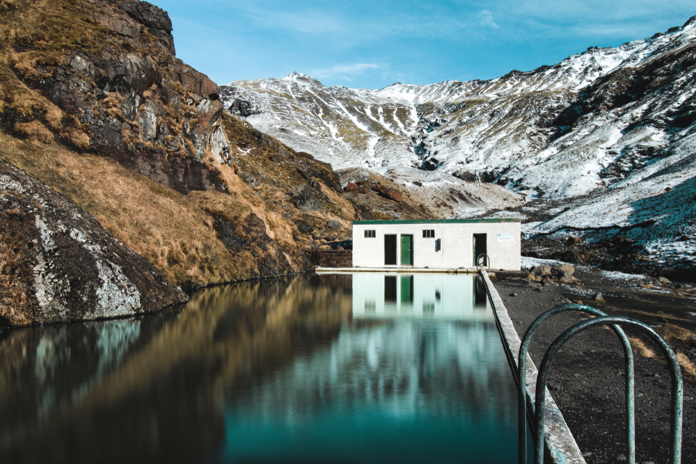 Seljavallalaug Hot Springs surrounded by mountains some with snow on them.  There is a green and white building