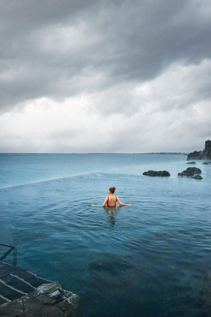 A girl in red swimsuit with manmade rocks with steam coming over the infinity edge