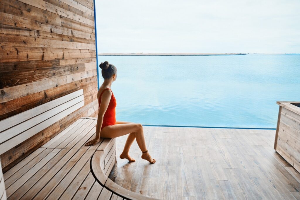 A girl in red swim suit in a wooden sauna overlooking the Atlantic Ocean with a glass window