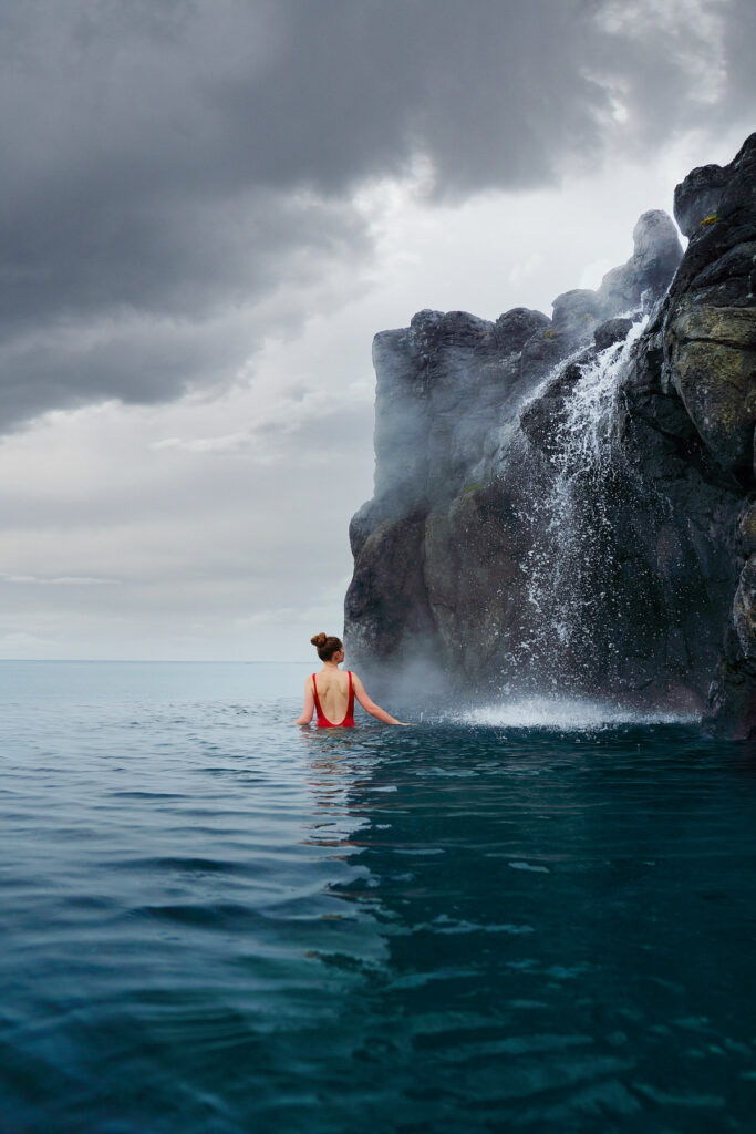 A girl in red bathing suit with her back towards a waterfall at the Sky Lagoon Iceland
