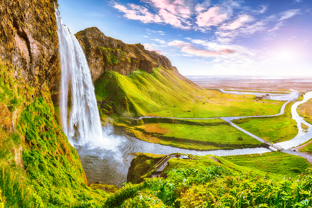 A slight aerial view of Seljalandsfoss waterfall from the side of the front of the falls. You can see the river that leads away from the falls, the walkway, and the cliffs.