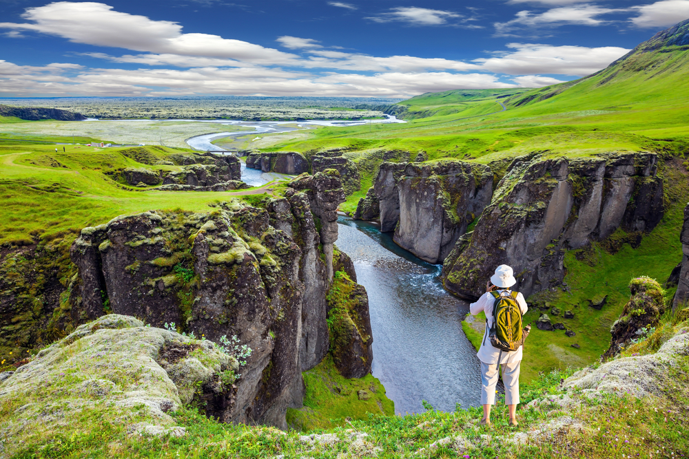 A person stands between a two riders which are green and alive with the summer in Iceland. This person takes a photo of this greenery in their white shirt and hat, with their back to the camera.