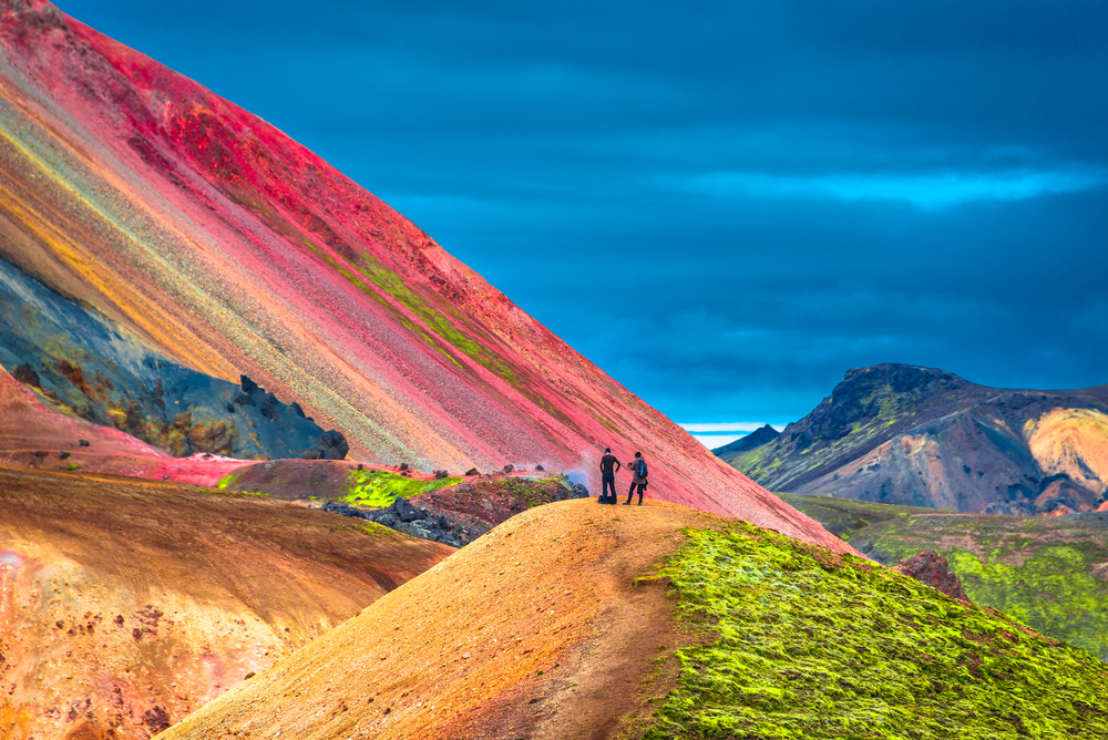 Hikers on their tour of the highlands stand and catch their breath with their back to the camera: they face the pink colors of the nearby mountain, thanks to the openness of the highlands during summer in Iceland.