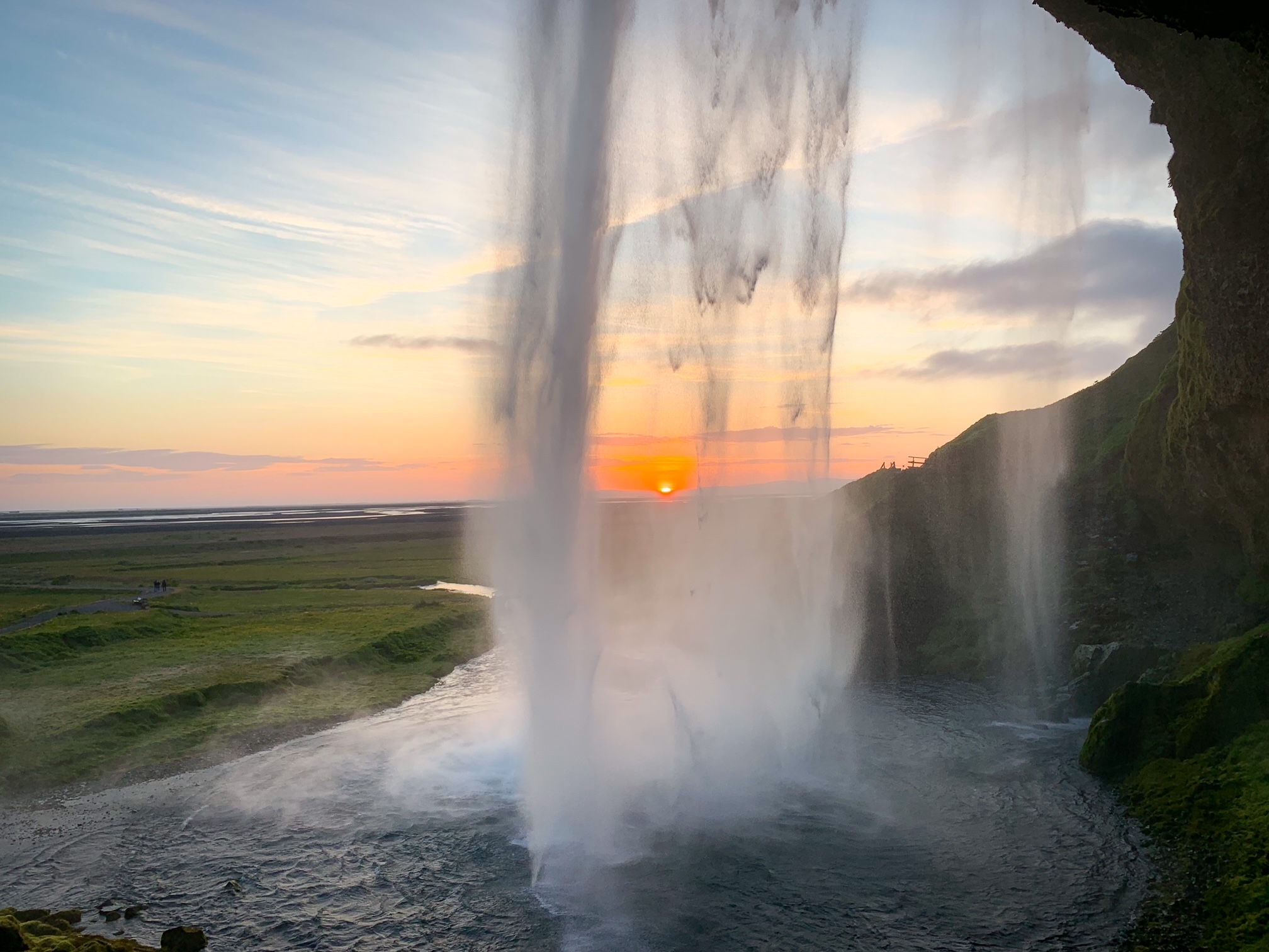 The view from behind the falls as the sun is setting on the horizon in the distance. Beyond the falls all you can see is miles of green fields. 