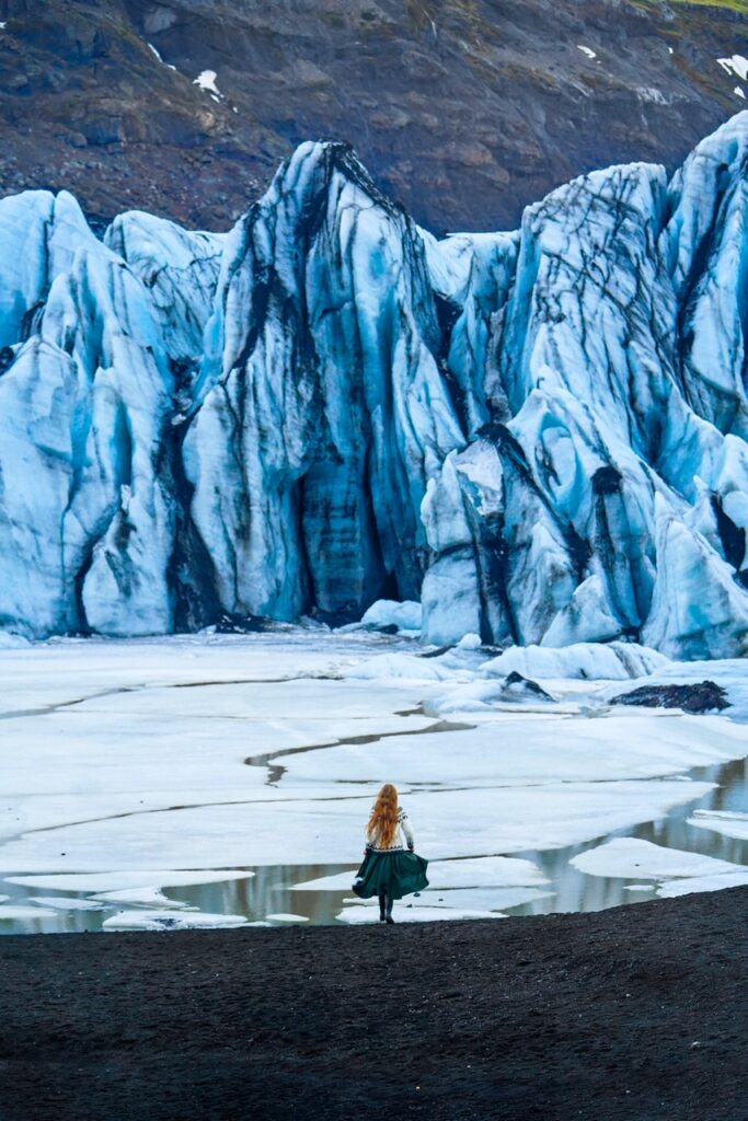 Girl with flowy skirt runs toward the glacier lagoon and the tall blue and black ice of the glaciers