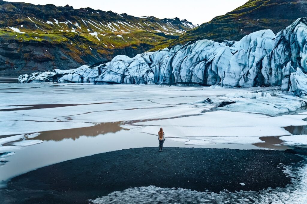 Girl standing in front of blue and black Solheimajokull glacier lagoonon a black sand beach.