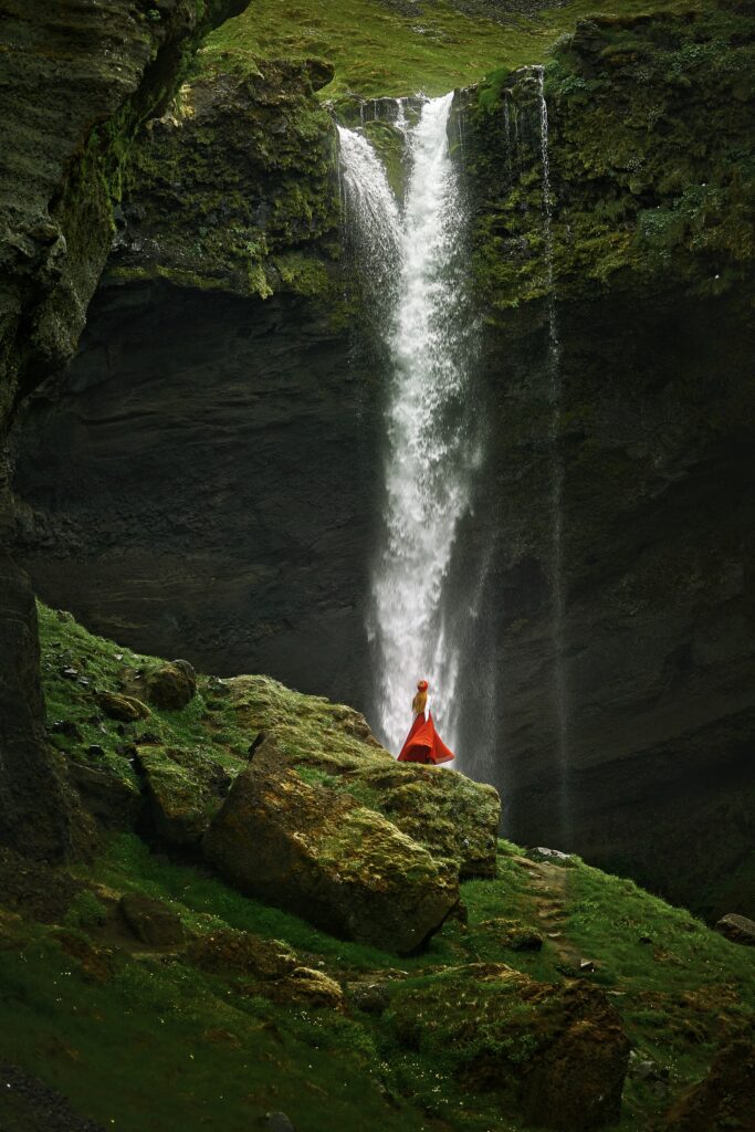 a woman in a long red skirt and matching hat standing on the hill next to Kvernufoss waterfall