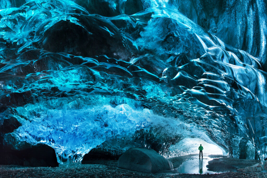 Person stands in opening of Sapphire blue ice caves in Iceland and look like perfect glassy scultpures 