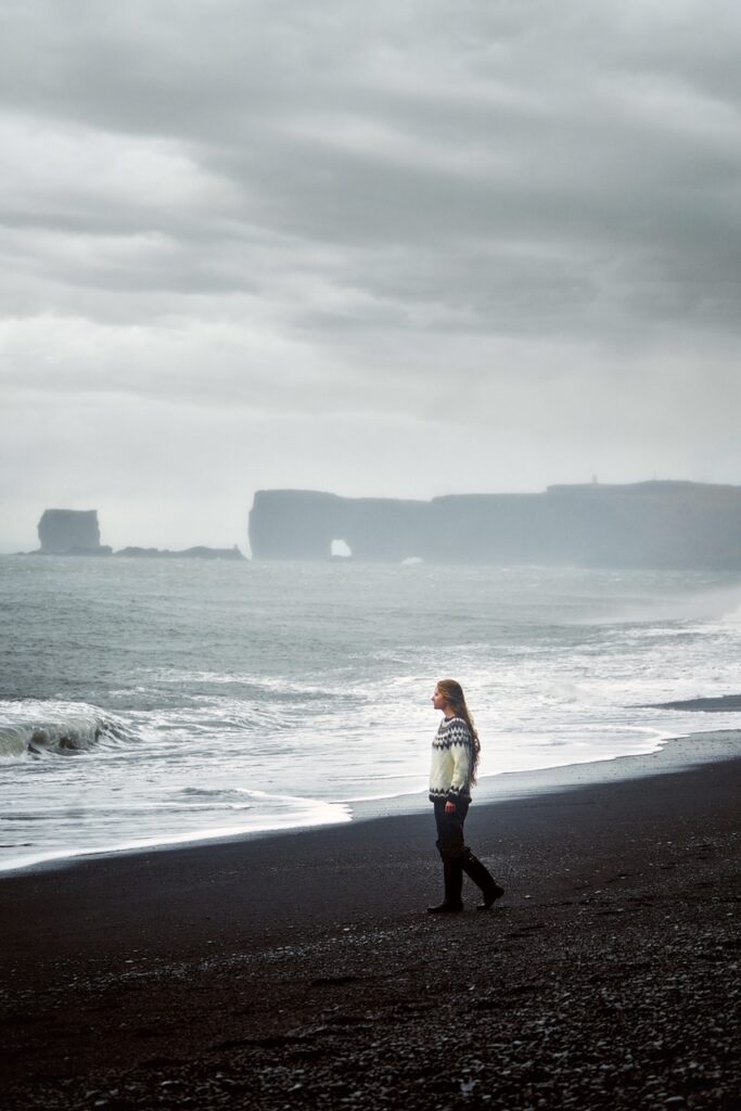 Girl standing by the black sand shore with Dyrholaey arch and lighthouse in the background is the perfect attraction near Vik