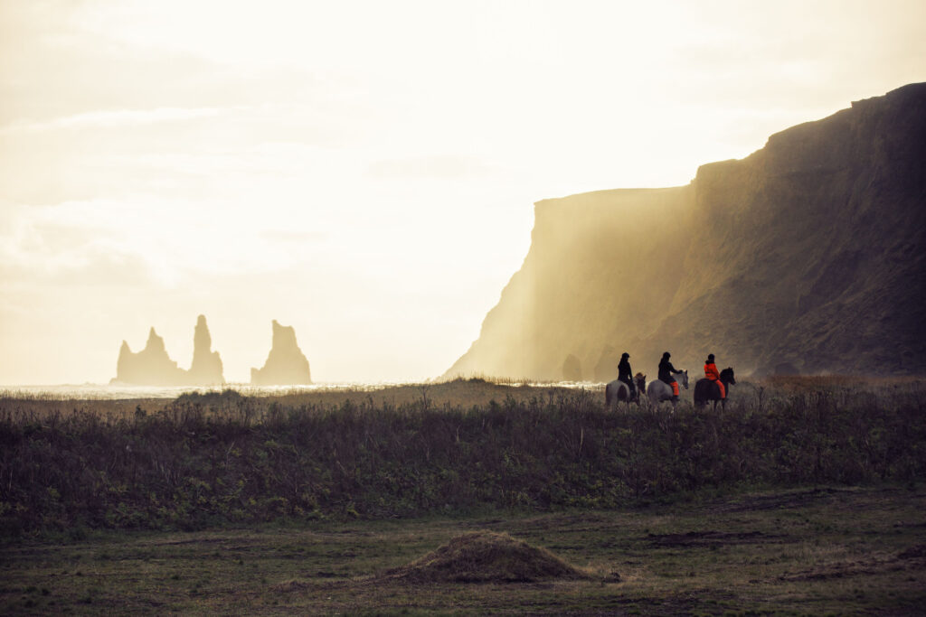 Three people horseback riding on black sand beaches near giant sea stacks during sunset is the one of the best things to do in Vik Beach
