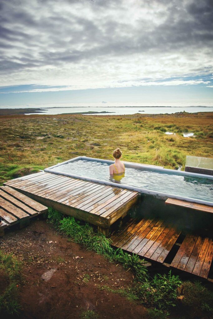 a woman sitting in a small hot spring in the middle of nature looking out at the ocean beyond