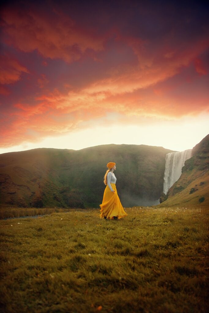 a woman in a yellow hat white shirt an long yellow skirt walking at sunset past Skogafoss waterfall in Iceland in April