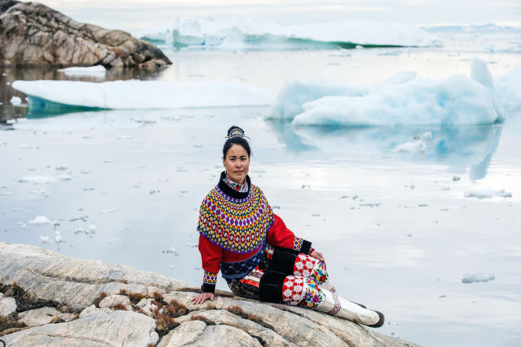 Native Greenland woman sits by icebergs displaying colorful Inuit geometric patterned dress in Greenland vs Iceland 
