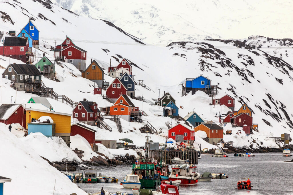Colorful houses line snow covered mountain by the bay where multiple fishing boats are seen in the early morning light