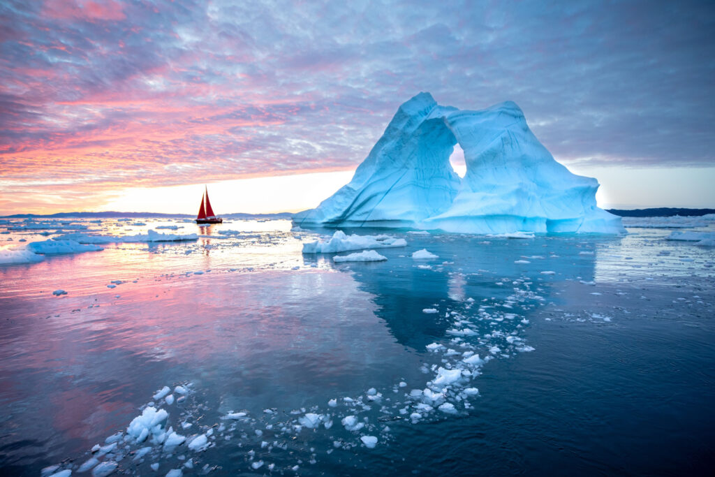Sunrises with pink shooting across the cloud covered sky while a sailboat drifts around a lazy blue iceberg