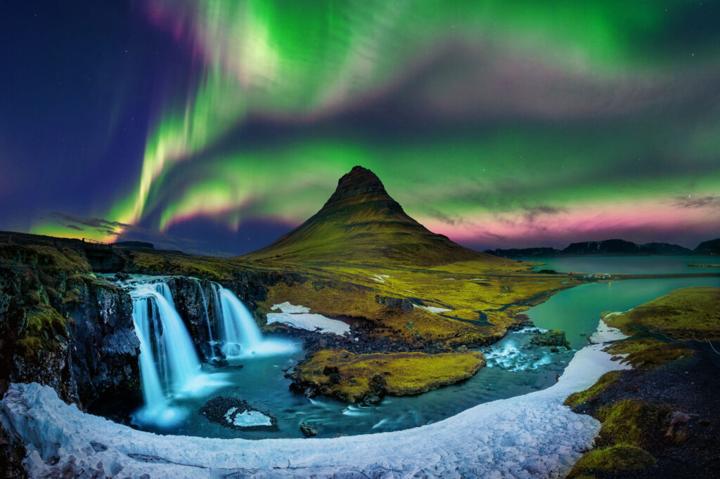 Colorful Northern Lights shine brightly illuminating a large Icelandic waterfall with moss covered mountain in the distance