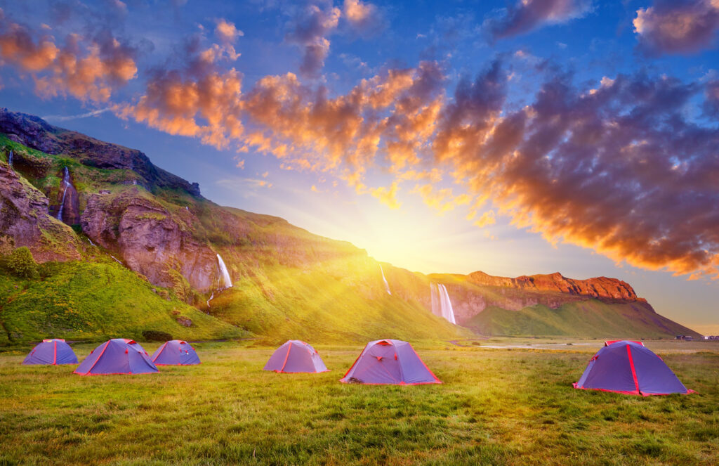 Dispersed tents at Iceland campsites with colorful sunrise peeping out over mountainside with many flowing waterfalls