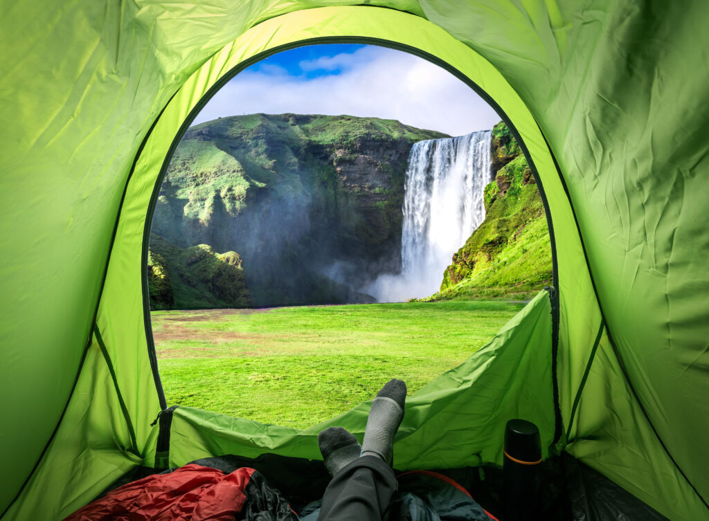 Person at iceland campsites with green tent, looking out at giant waterfall and mossy mountains