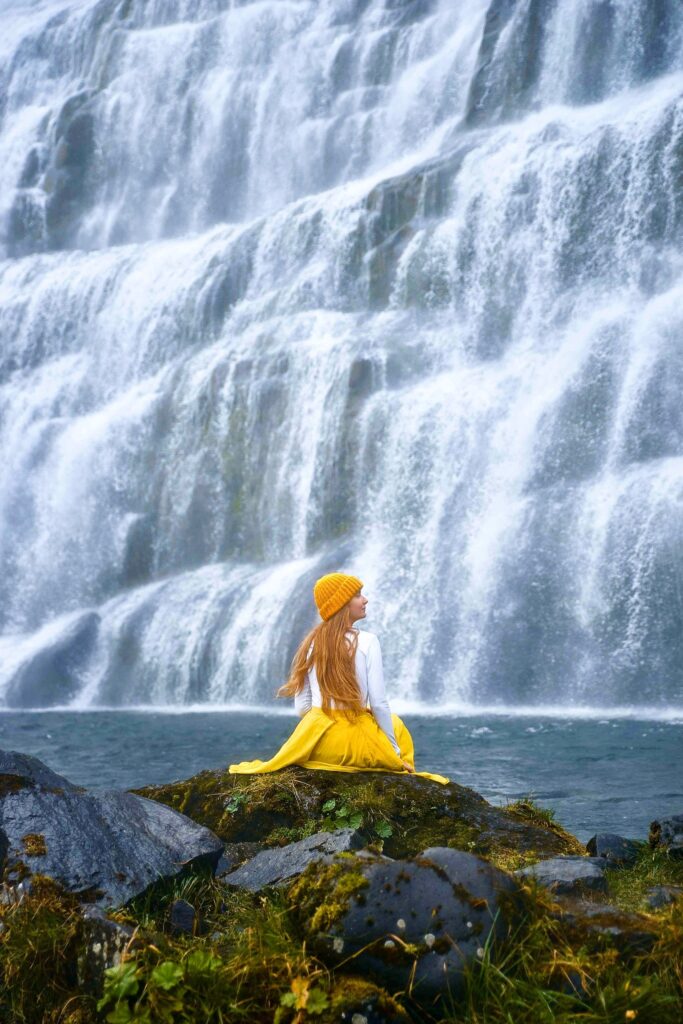 Girl in yellow skirt sits before monumental cascading waterfalls for awesome photos of Iceland
