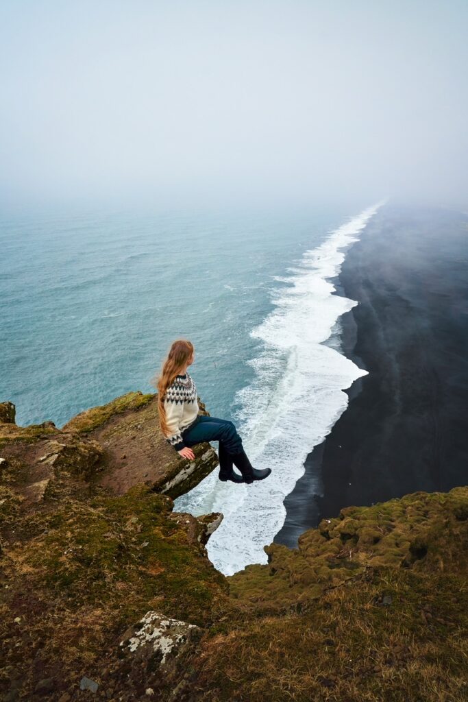 Girl sitting on a rock edge, high above black sand beaches where violent white-capped waves of the Atlantic approach the shoreline at one of the best spots for Iceland photos.