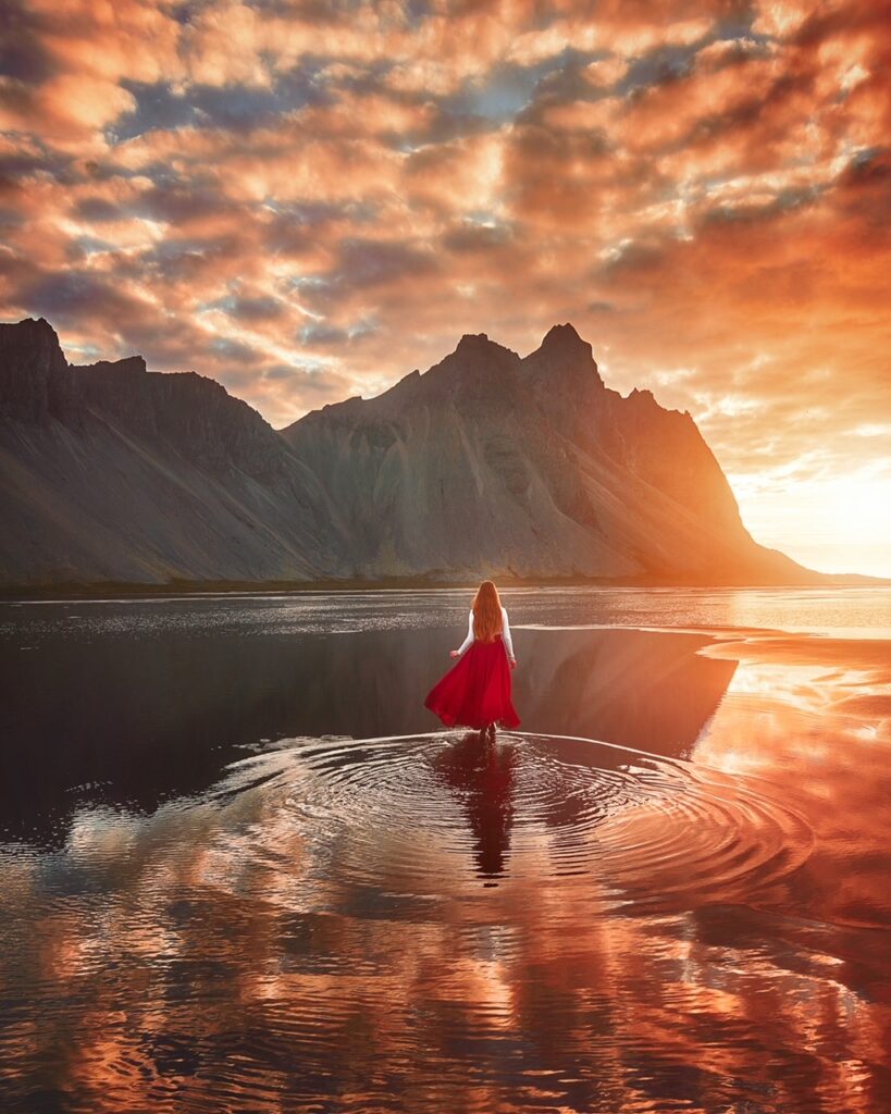 Girl in red skirt walks toward large mountain range as if she is walking on water under a gorgeous orange sunrise.
