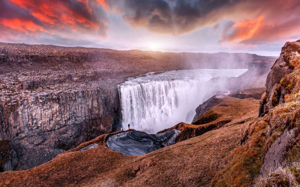 Couple stands beside a small mountainside pool looking at a colorful sunset over powerful waterfall.