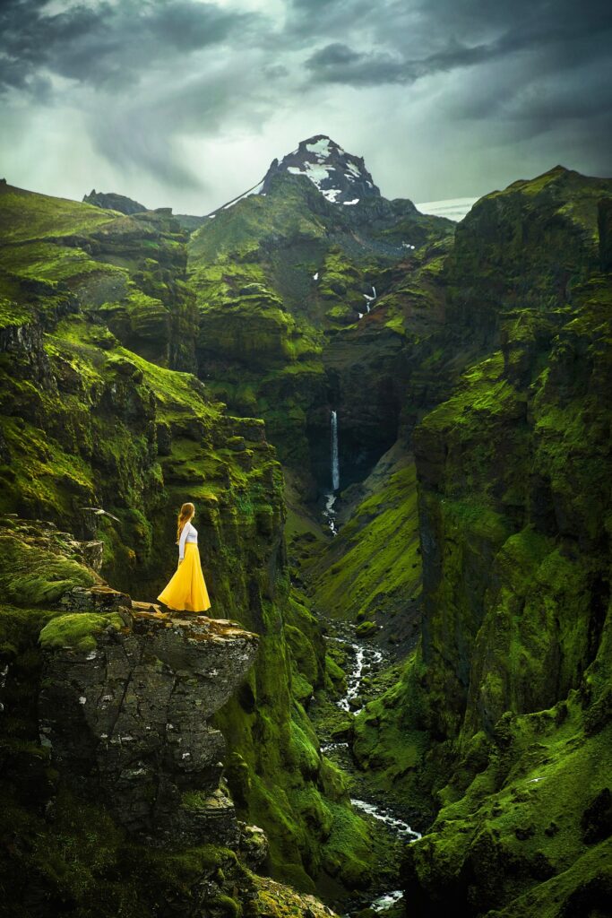 Instagrammable spot in Iceland with a girl standing in dress beside mossy covered green canyon walls with a distant waterfall hanging in front of a distant craggy mountain.