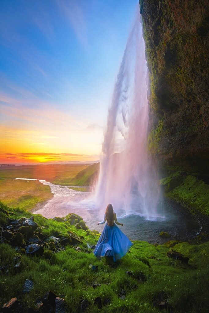 Instagrammable spot in Iceland shows a girl in blue skirt runs toward waterfall that you can walk behind as the sun illuminates the sky with a firey orange