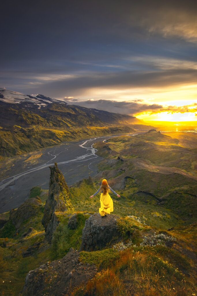 Girl in yellow flowing skirt dances on mountaintop which over looks a valley with a river flowing through it in gorgeous Iceland photography