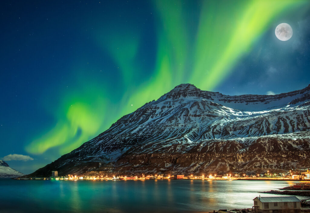 Snow covered mountain with a tiny lit up town casting warm lights across the water and green northern light and a full moon 

