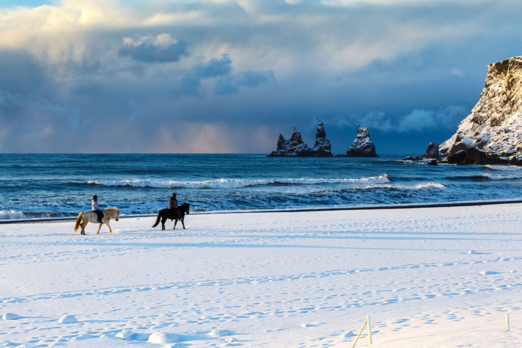 Two people on the best horseback riding tours in South Iceland on a snowy Oceanside with towering sea stacks in the distance