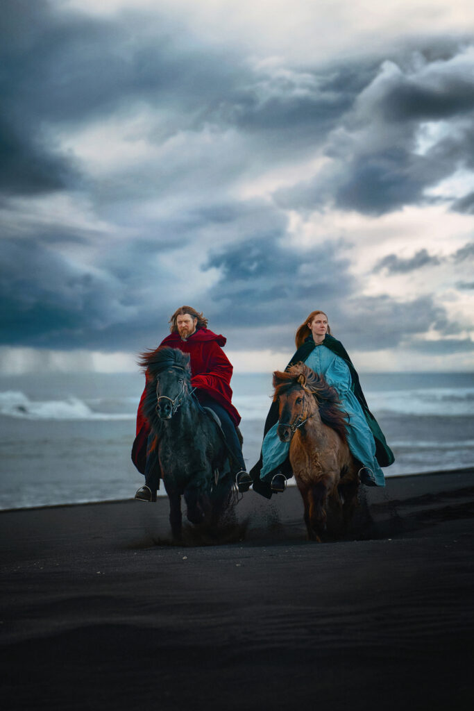 Viking man and woman on horseback riding tour in Iceland on a stormy day along the black sand beach
