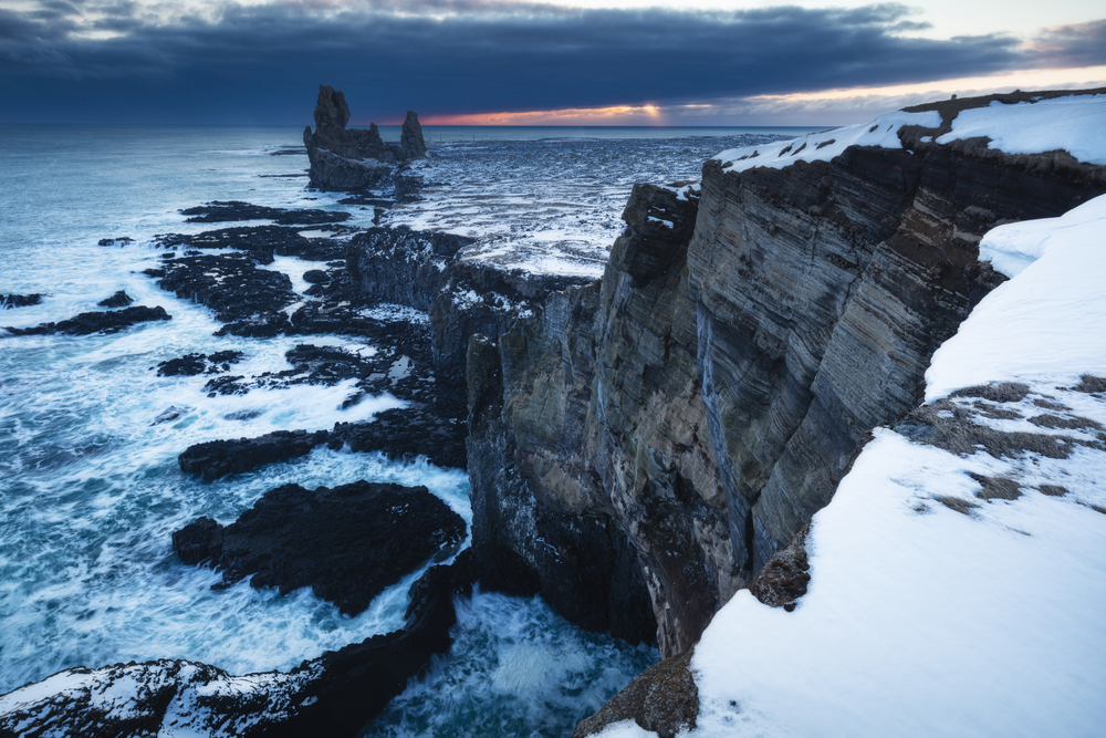 Londrangar in the distance with a snowy rock wall in the foreground and the Atlantic Ocean crashing up against the rocks