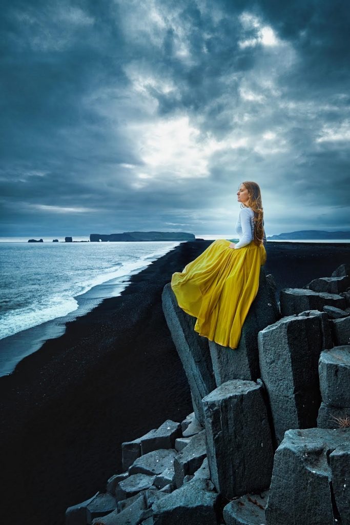 A woman in a yellow skirt sitting on a basalt column overlooking Reynisfjara Black Sand Beach.