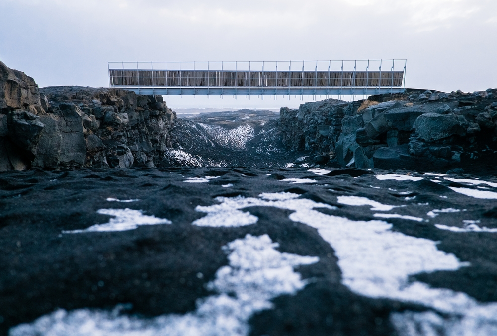 a look at the bridge between two continents from the slightly snow covered black sand beach between the bridge with lava rock on the sides of the bridge and mountains in the background
