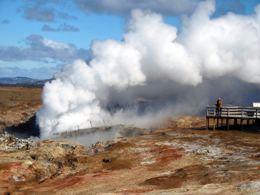 two tourists standing on a raised platform looking at the smoke billowing from the Gunnuhver hot springs surrounded by a brown and red landscape