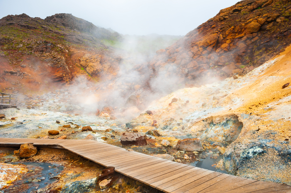 one of the boardwalks at Seltun geothermal area with steam rising beside it as well as boiling mud pots and yellow and brown mineral deposits