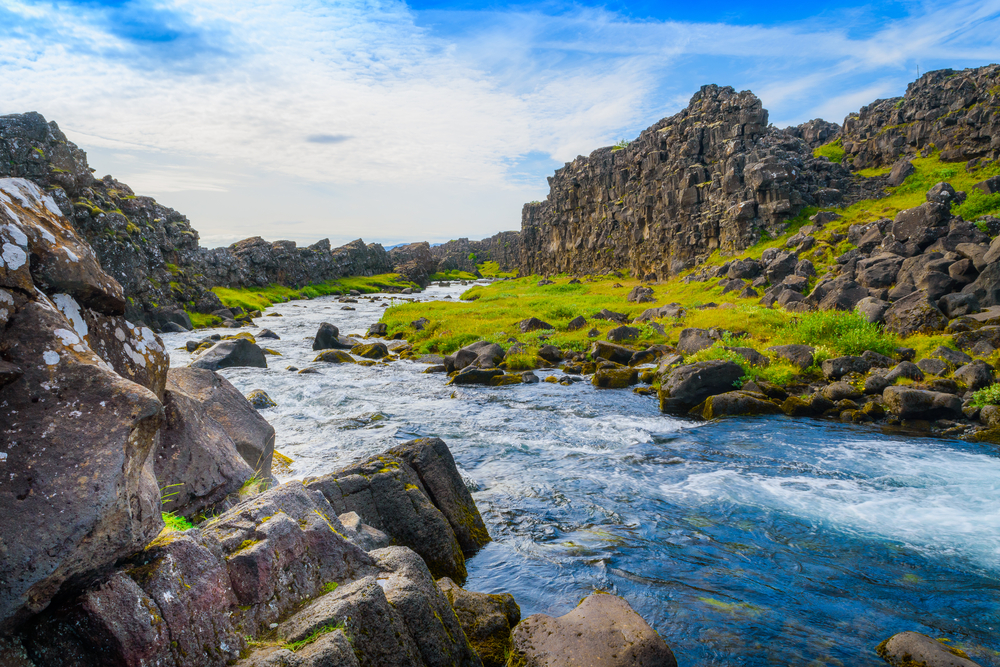 Water downstream from a waterfall in Thingvellir National Park in Iceland. There are rocks on the riverbank and grass. 