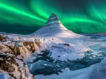 Green northern lights over snow-capped Kirkjufell and Kirkjufellfoss waterfall in Iceland in February.