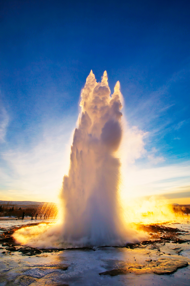 The Strokkur Geyser gushing high in the air with the sun shining from behind it.